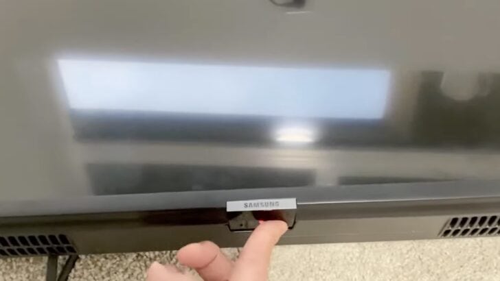 Photo of person turning on samsung tv using the power button under the samsung logo in the middle underside of the tv